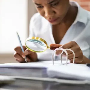 A woman auditor analyzes a binder of documents with a magnifying glass